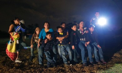 Central American children in a group looking scared at night next to an immigration officer with a flashlight.